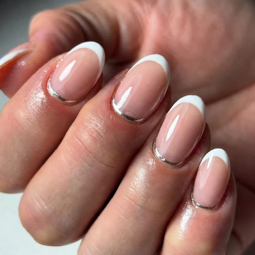 Close-up of reverse French tip nails with a nude base, metallic cuticle line, and white curved tips on almond-shaped nails.
