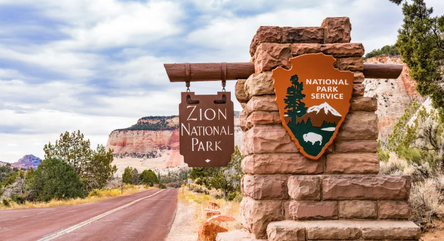Zion National Park entrance sign with red rock cliffs and scenic road in southern Utah.
