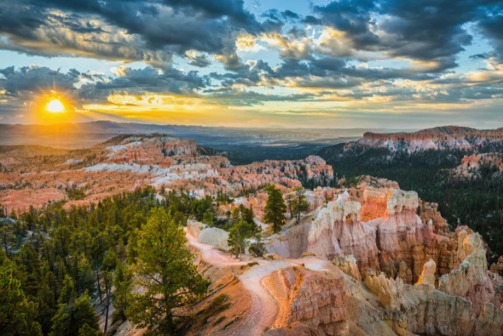 Bryce Canyon National Park at sunrise with hoodoos, pine trees, and dramatic clouds over red rock formations.