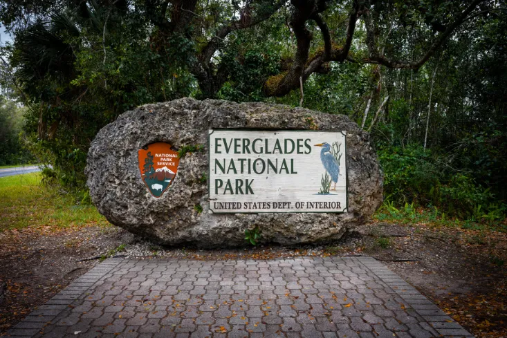 Stone entrance sign for Everglades National Park with park emblem and heron illustration, surrounded by dense forest in Florida.