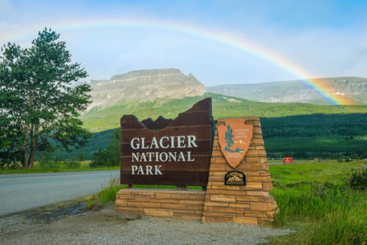 Glacier National Park entrance sign with rainbow in the background, mountain view, and National Park Service emblem.