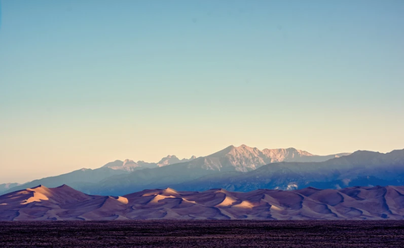 Golden sand dunes at Great Sand Dunes National Park with mountain backdrop at sunrise under a clear sky.