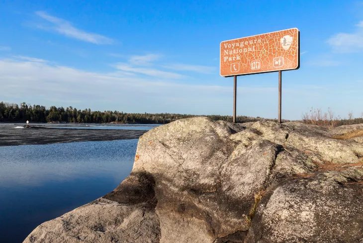 Voyageurs National Park sign on a rock overlooking a calm lake under a clear blue sky in Minnesota.